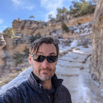 Selfie of Zachary Crockett on an icy path inside the Grand Canyon, sunglasses, black puffy jacket, blue sky, tan cliffs, small pine trees